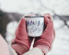 Pink mittened hands holding a steaming hot beverage in The Everyday Green's enameled camping mug, featuring the Watercolor Forest designed personalized with your name in brush script over a blue and purple watercolor pine forest.