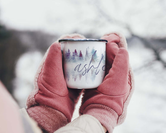 Pink mittened hands holding a steaming hot beverage in The Everyday Green's enameled camping mug, featuring the Watercolor Forest designed personalized with your name in brush script over a blue and purple watercolor pine forest.